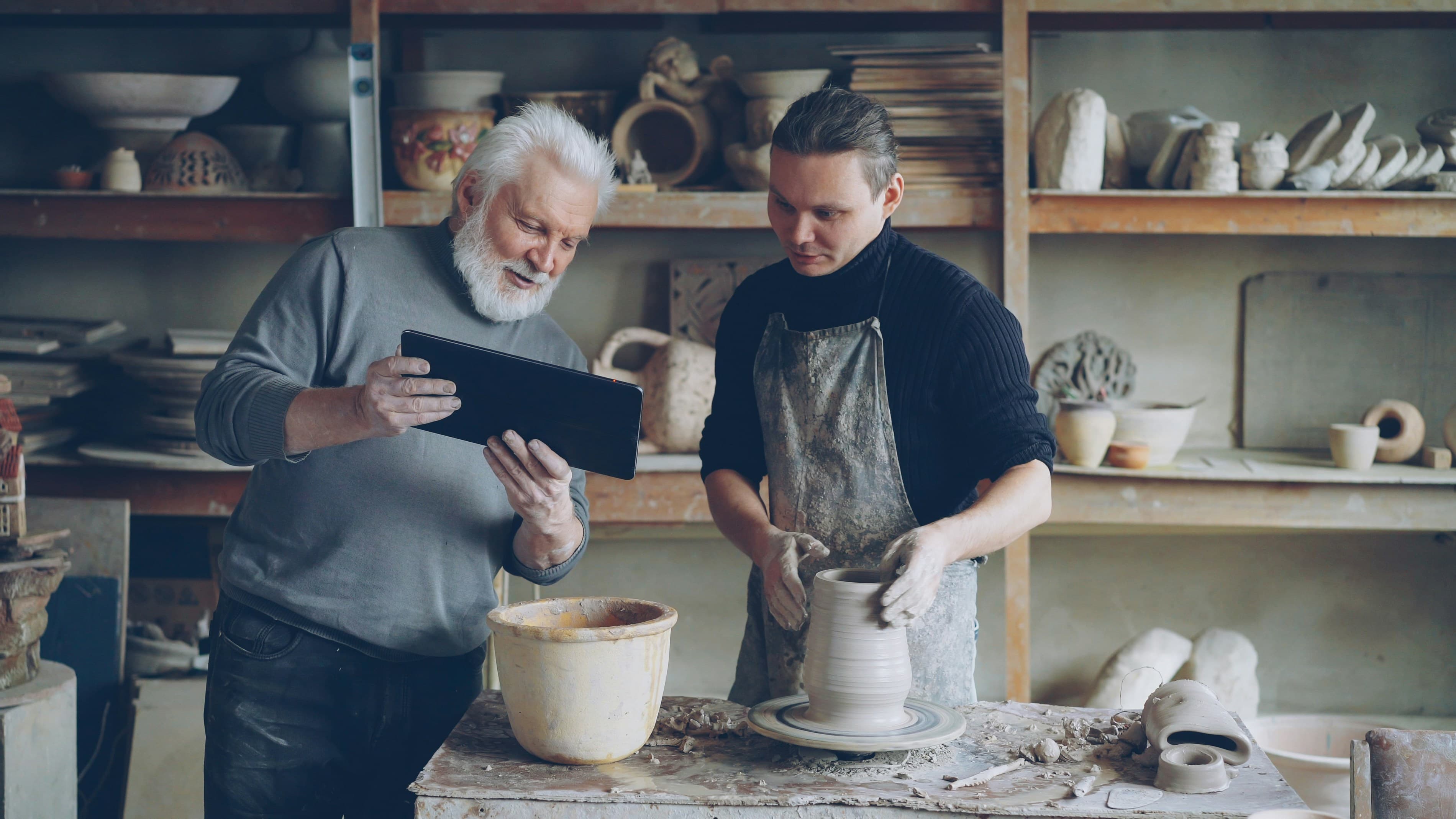 Two generations of small business owners reviewing information on a tablet in a pottery workshop.