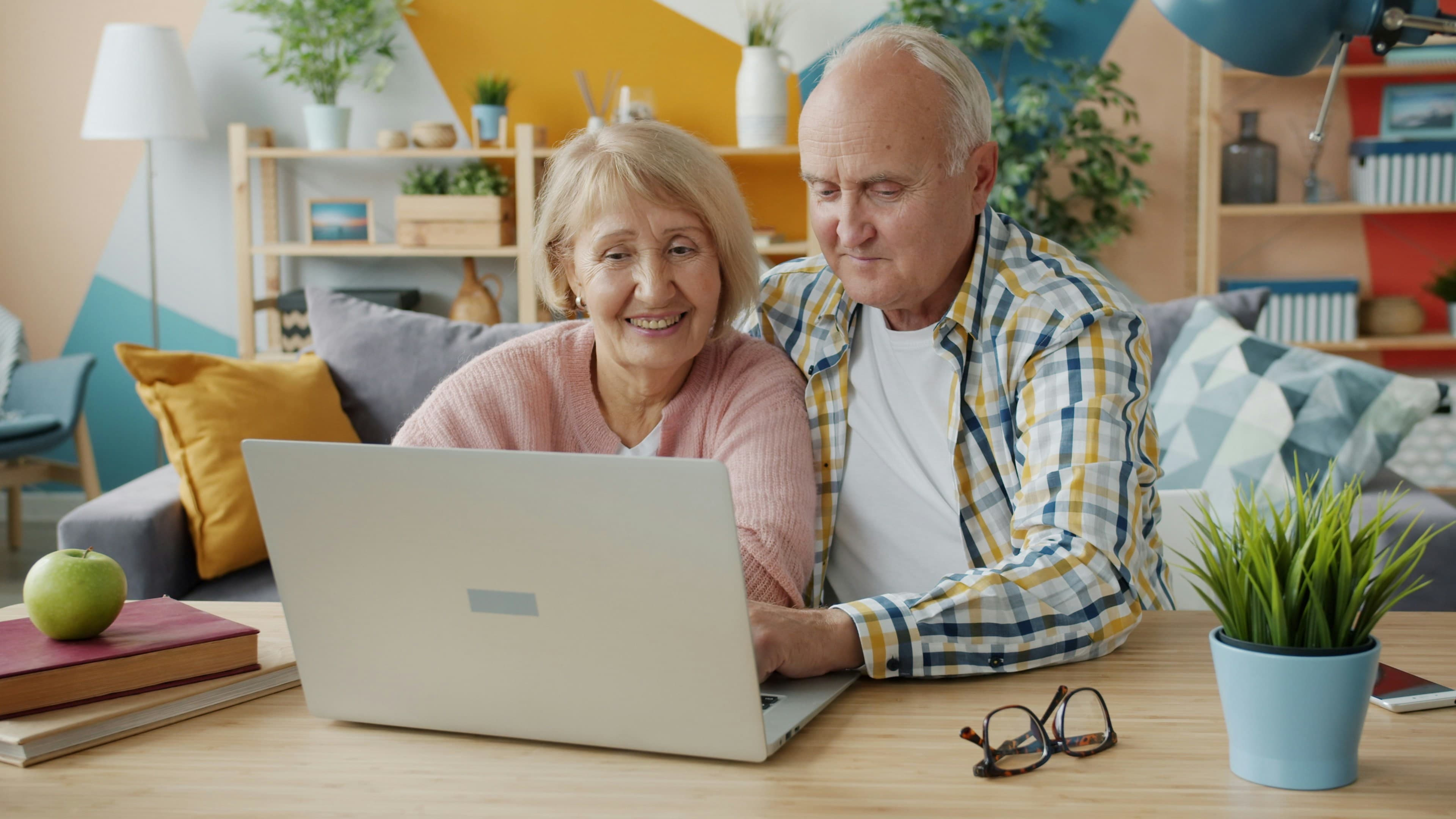 An elderly couple smiling while looking at a laptop together at home, sitting comfortably in a bright and colorful living room.