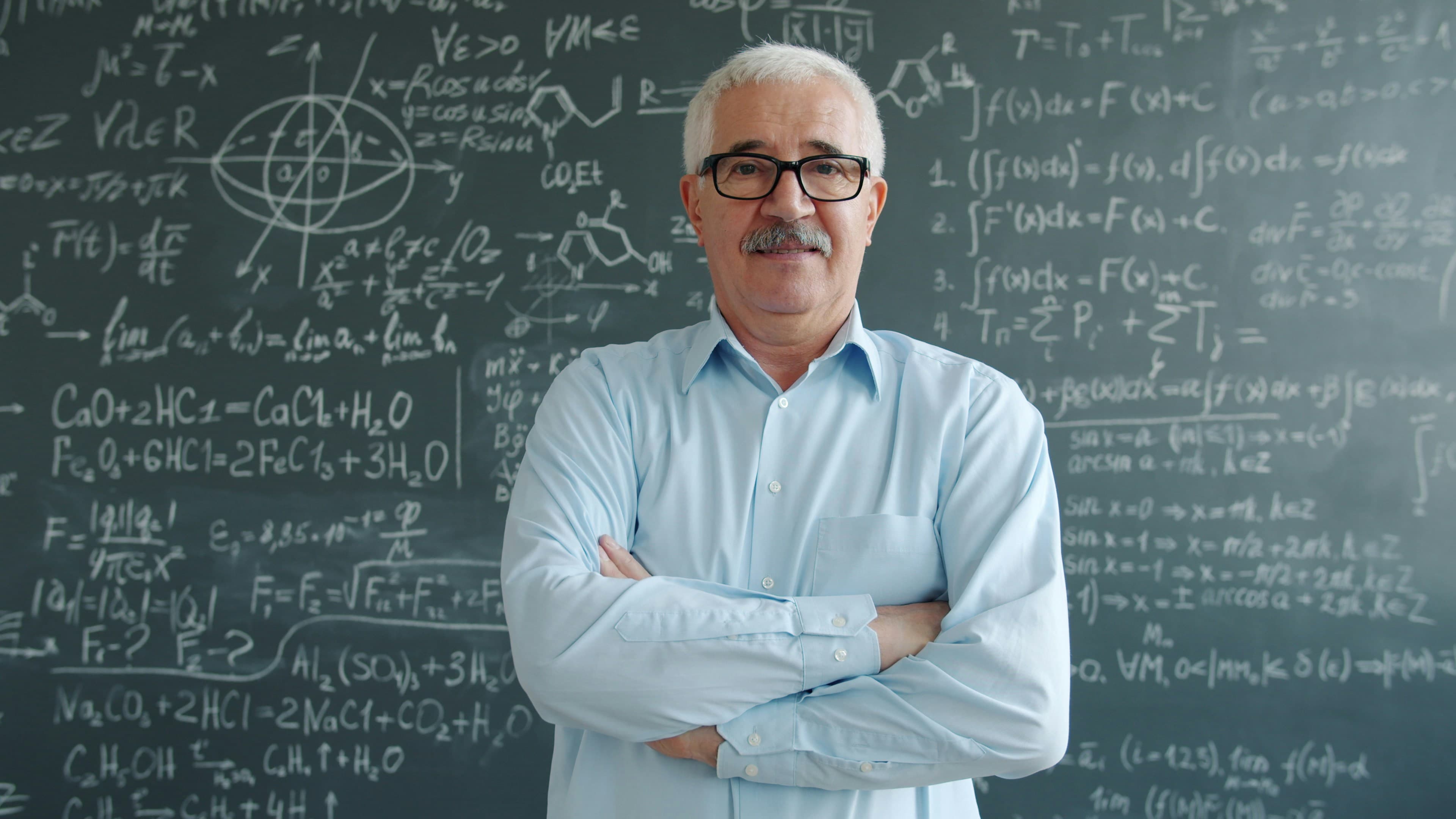 A smiling older male teacher with glasses standing with his arms crossed in front of a chalkboard filled with mathematical equations.